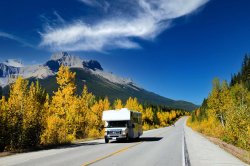Motorhome driving along Icefields Parkway, Canada with mountains and trees in background and blue sky.