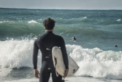 Man stands in foreground with surfboard, looking out to the waves in front of him